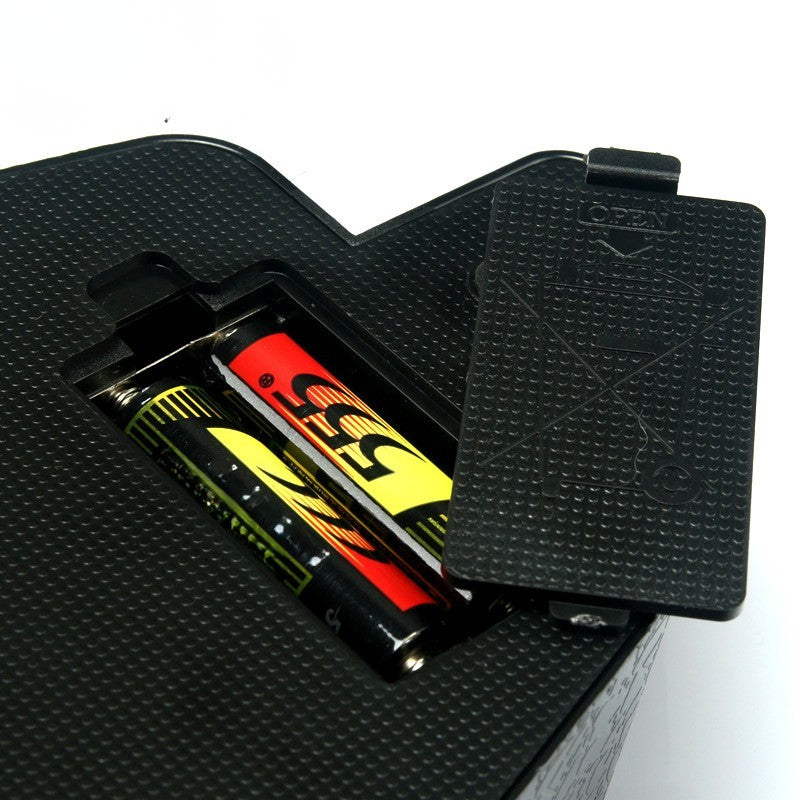 A close-up of a black textured battery compartment cover with its lid propped open, showing two batteries inserted inside. The batteries are black with red and yellow graphics and the text '555'.