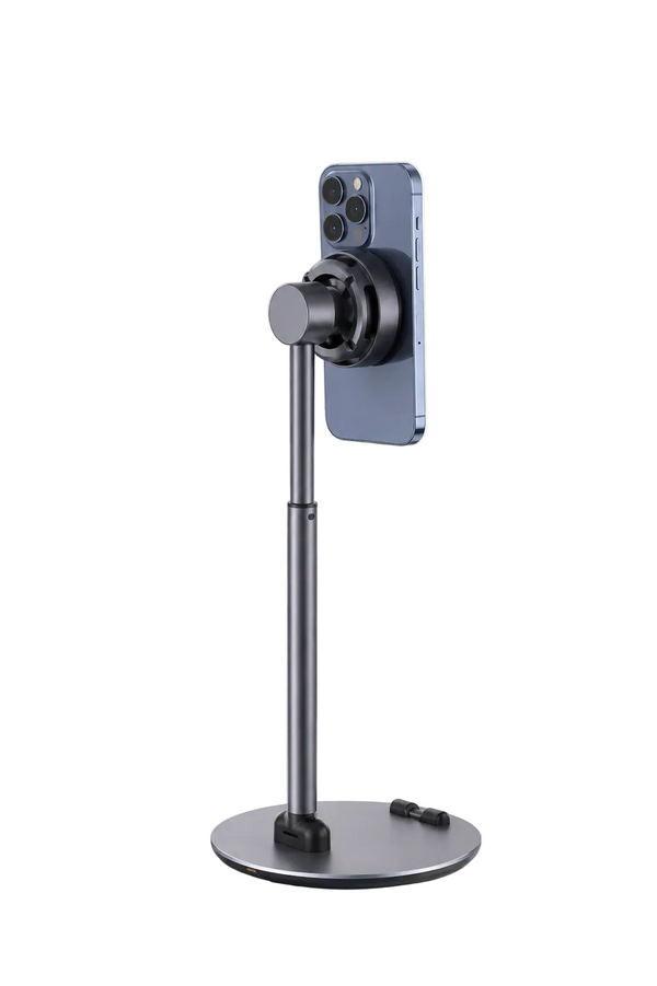 A dark gray adjustable smartphone stand holding a blue-gray iPhone, set on a light marble tabletop with a blurred potted plant and books in the background.